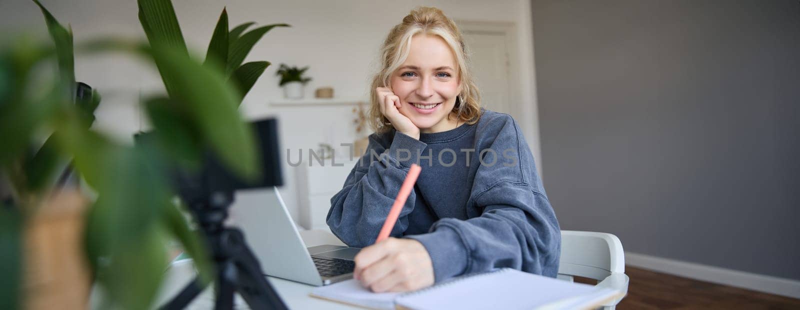 Cute smiling girl sits in a room, writes down notes, doing homework, records video of herself on digital camera, creates content for vlog, lifestyle blogger doing daily routine episode.