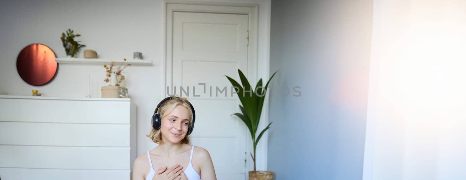 Vertical shot of young sporty woman in headphones, sits on rubber mat after workout, holding hands on chest, looks aside with peaceful, relaxed face expression.