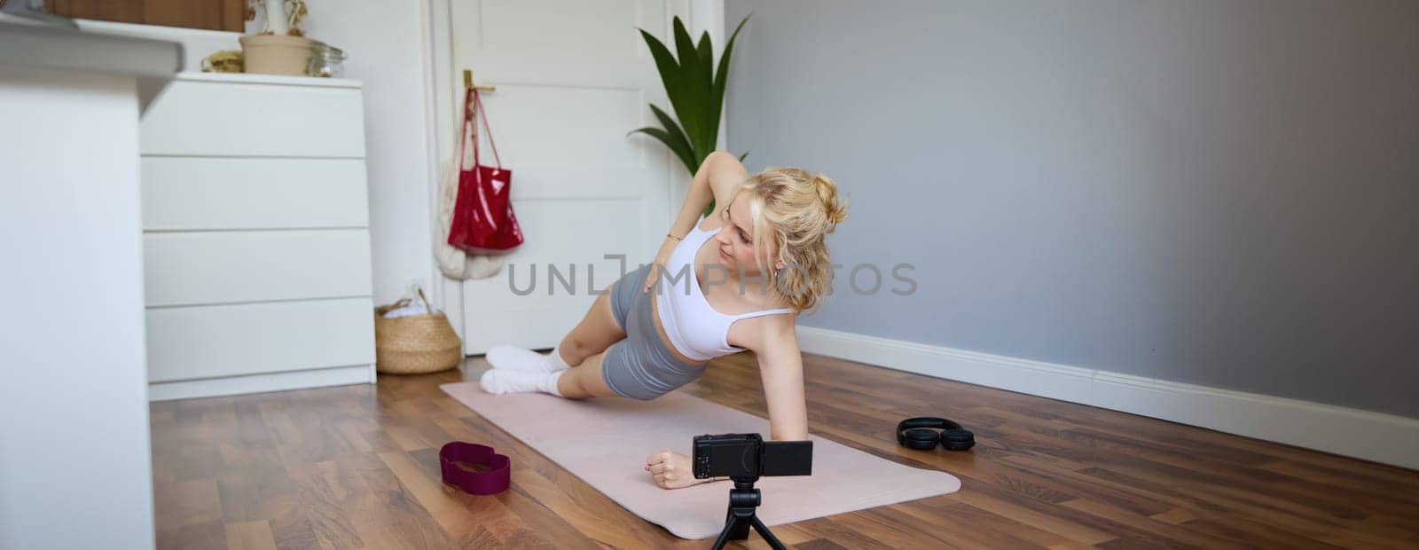 Portrait of young woman, fitness instructor recording video of her standing in a side plank, doing workout at home on camera, creating content for social media by Benzoix