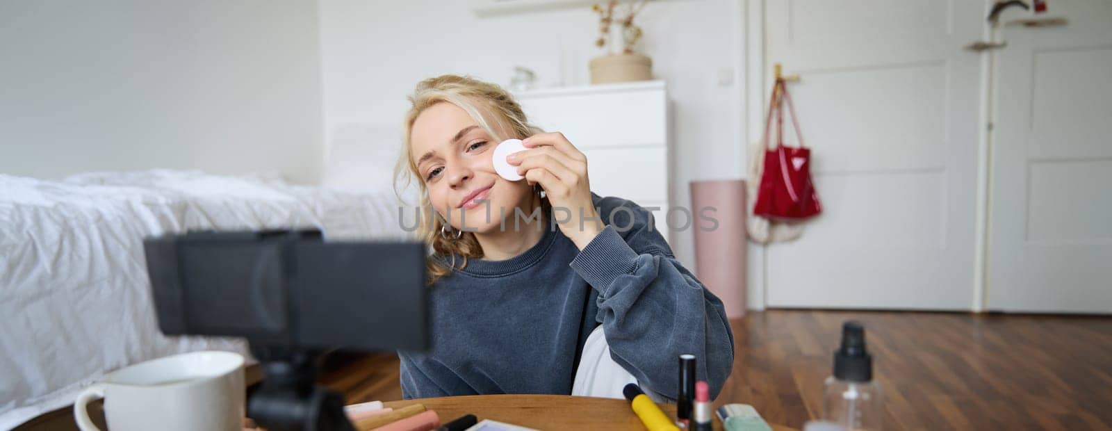 Portrait of smiling young woman applies makeup, shows beauty tutorial on video, records vlog on digital camera in her room, recommends cosmetic products.