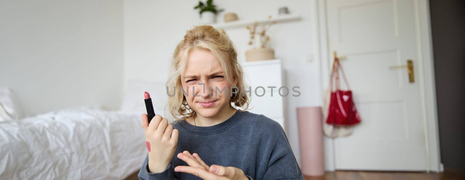 Portrait of young blogger, woman recording video about beauty, makeup products, showing lipstick and grimacing, express negative opinion, creating a lifestyle vlog content.