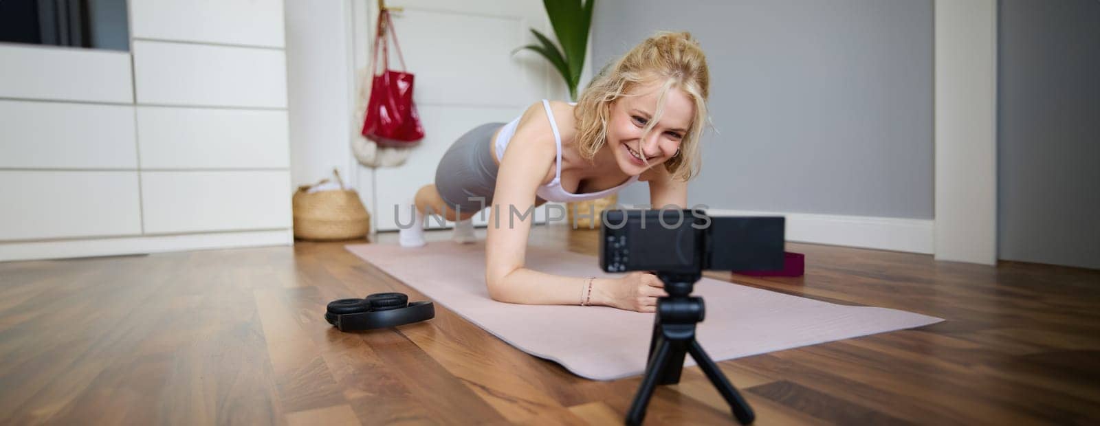 Portrait of young sporty vlogger, fitness instructor standing in plank on rubber yoga mat, recording video of herself doing workout at home by Benzoix