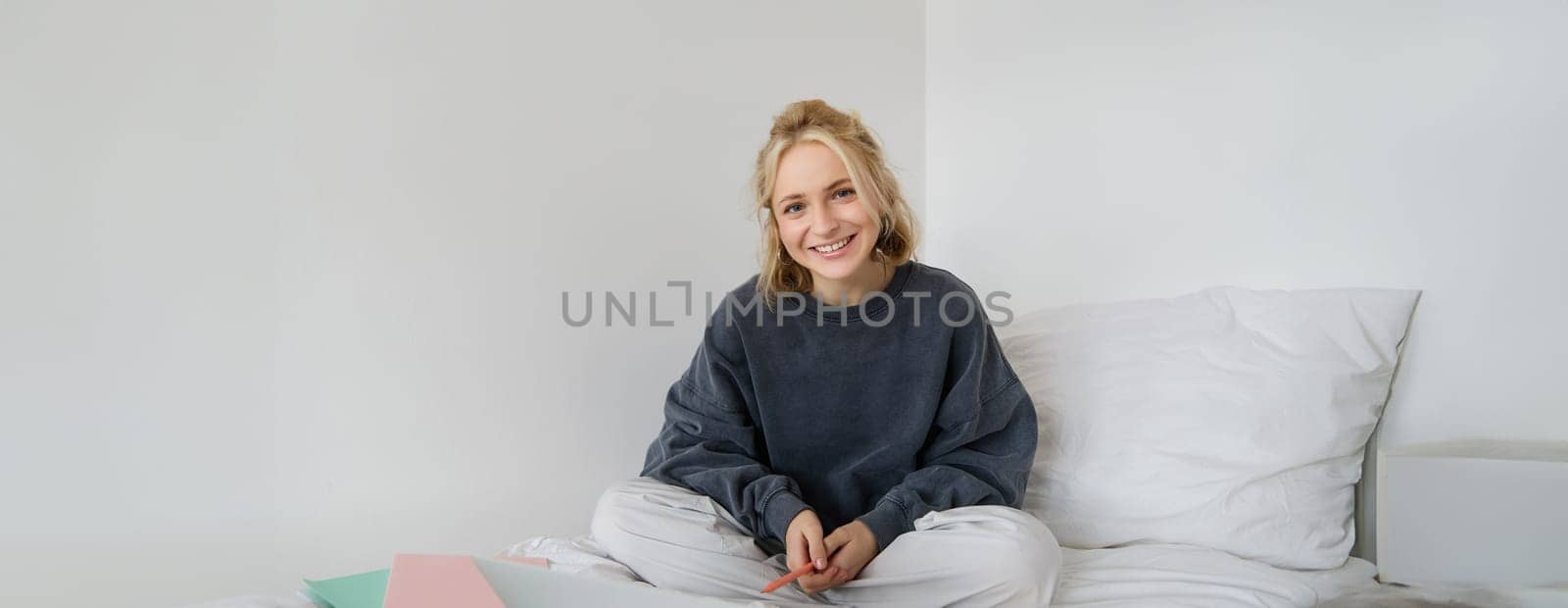 Portrait of young beautiful woman sitting on bed with laptop and notebooks, working from home, freelancing. Female student studying online, video chats, connects to lesson remotely.