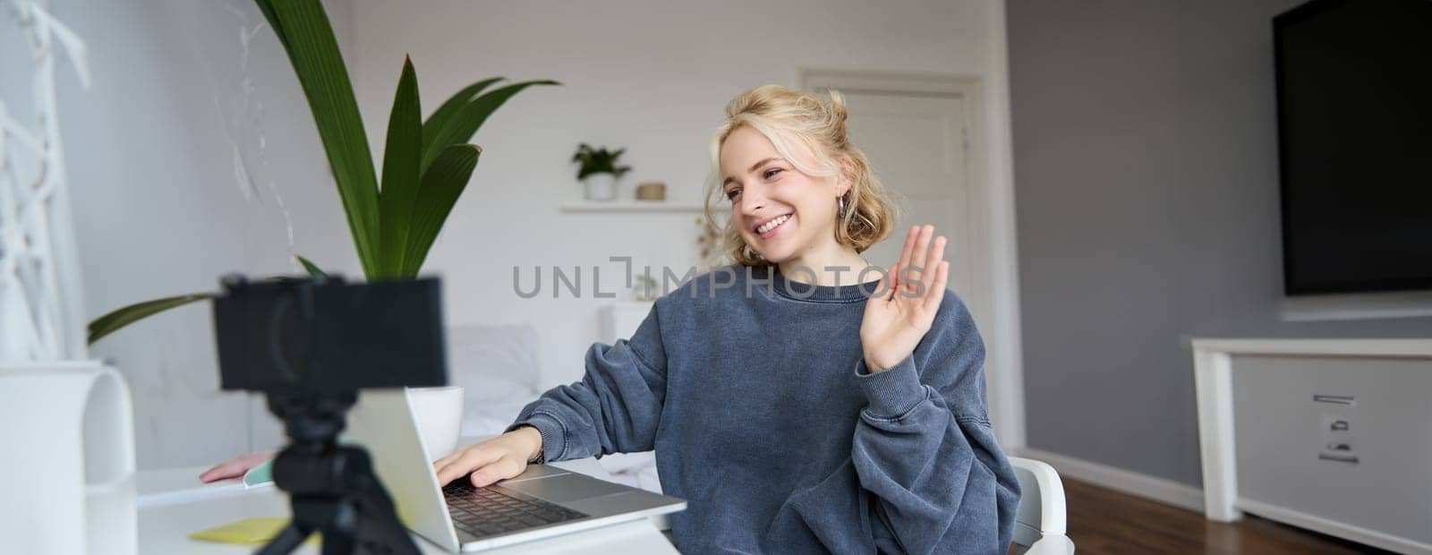 Image of smiling beautiful young woman, chats online on laptop, waves hand, says hello, records video on digital camera, vlogging, creates content for her blog by Benzoix