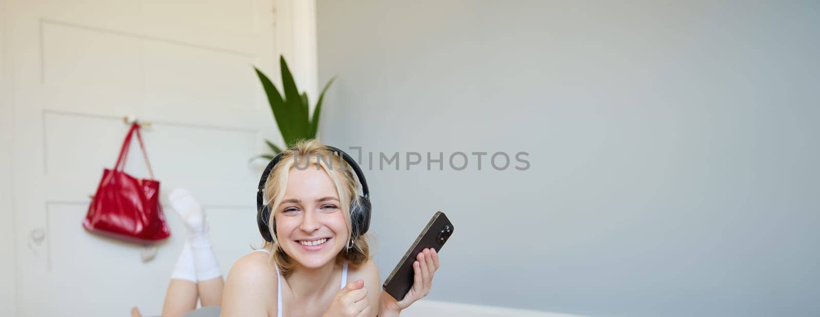 Vertical shot of young blond woman in headphones, lying on rubber mat, using laptop, holding mobile phone, listens to music or workout podcast, doing exercises at home by Benzoix