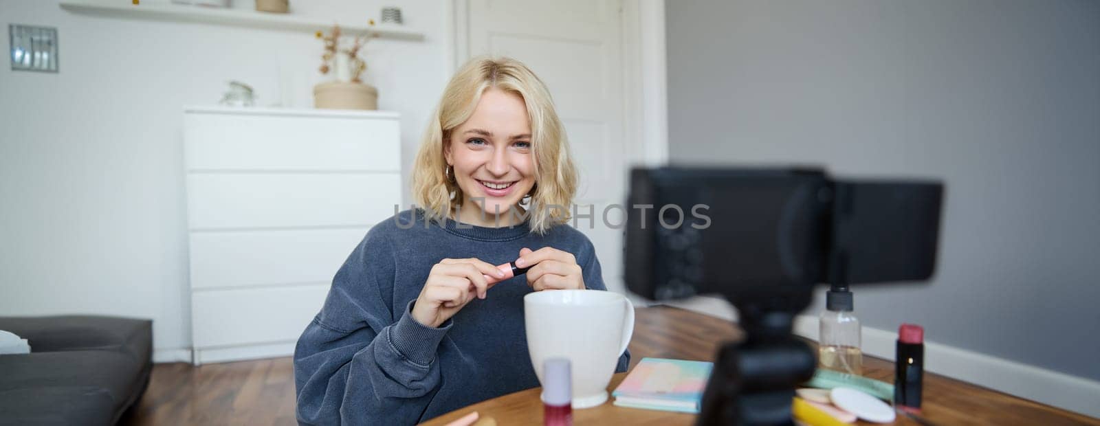 Portrait of blond smiling woman records a lifestyle blog, vlogger or makeup artist recording video for social media, holding mascara, reviewing beauty products for followers online by Benzoix