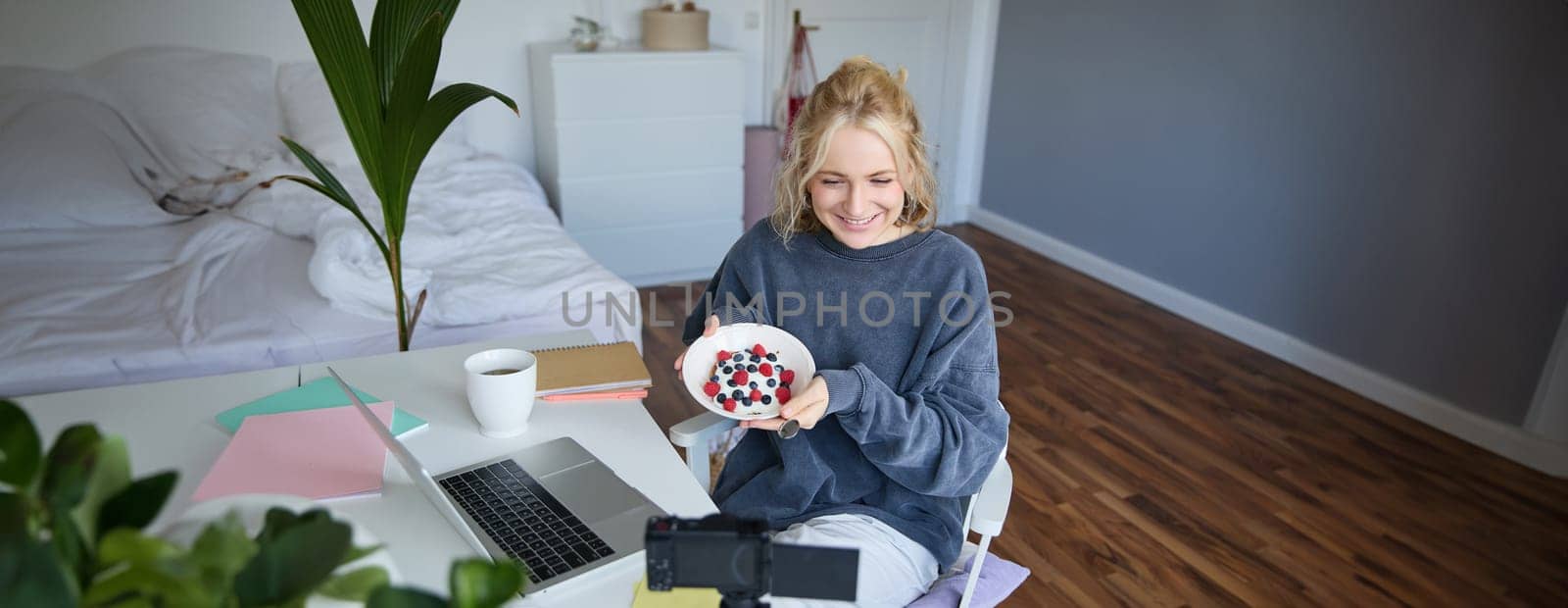 Portrait of cute young girl vlogger, showing her breakfast on camera, recording vlog in her bedroom, holding dessert, eating.