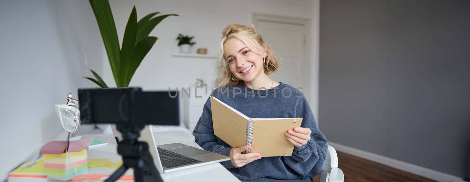 Portrait of cute young woman, teenage girl records video on digital camera, uses laptop to create lifestyle content, shows notebook, reads notes, using computer in her room.