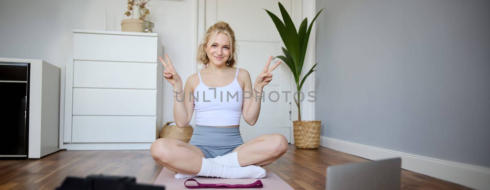 Portrait of cute blond woman shows peace sign, smiles, records yoga vlog on digital camera, workout at home on rubber mat in room by Benzoix