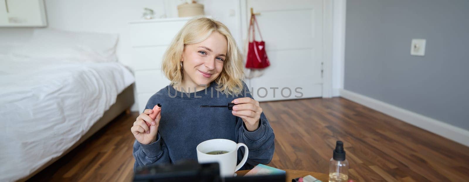 Portrait of cute lifestyle blogger sits in her room in front of video camera, recording for social media account, putting on makeup, reviewing beauty products, holding mascara.