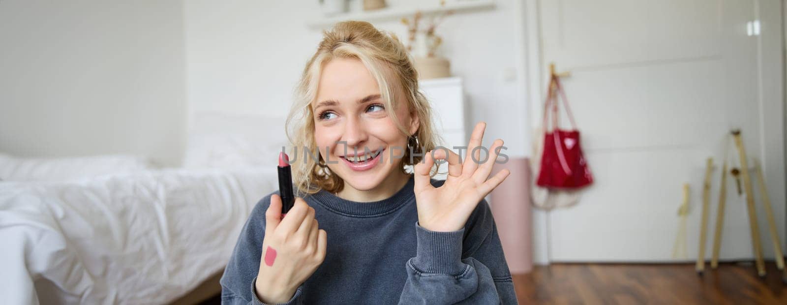 Young female blogger, content creator showing lipstick and okay hand sign, recommending beauty product for her audience on social media, recording vlog in room by Benzoix