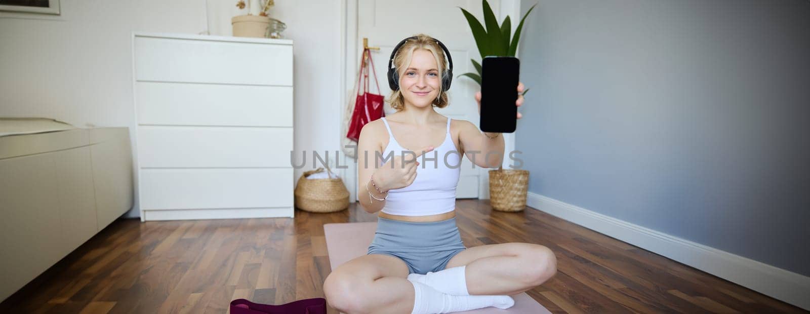 Portrait of smiling, fit and healthy, fitness instructor at home, pointing finger and showing her smartphone screen, recommending sports app for workout.