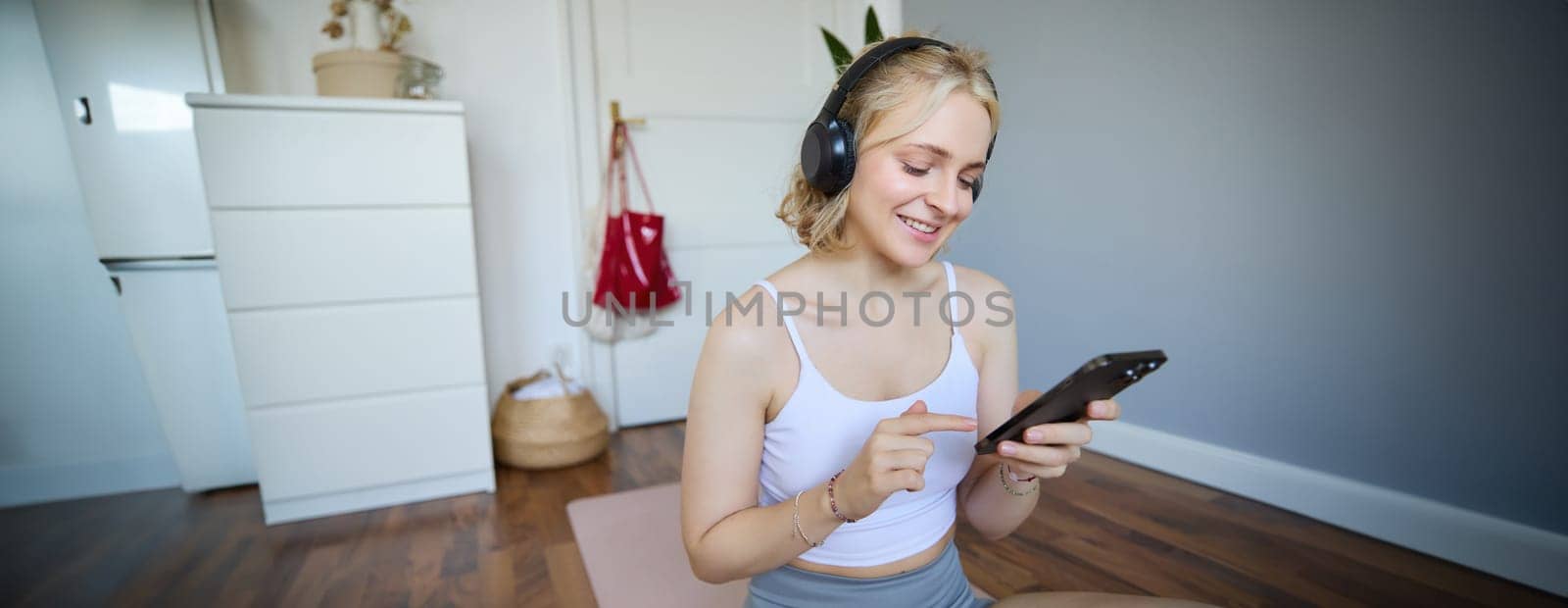 Close up portrait of blond young woman choosing fitness podcast, looking at her mobile phone, checking workout app on smartphone, wearing wireless headphones.