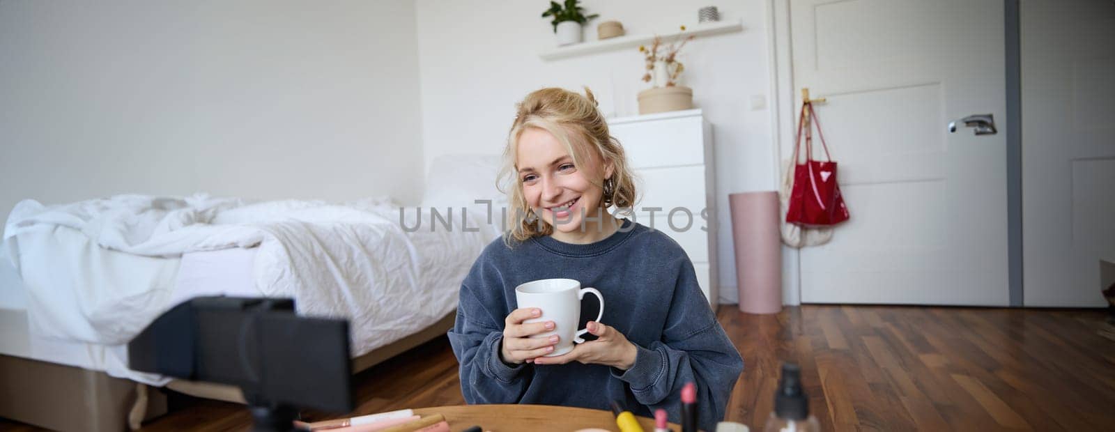 Portrait of beautiful social media beauty blogger, sitting in front of digital camera on floor in bedroom, drinking tea and chatting, talking to followers.