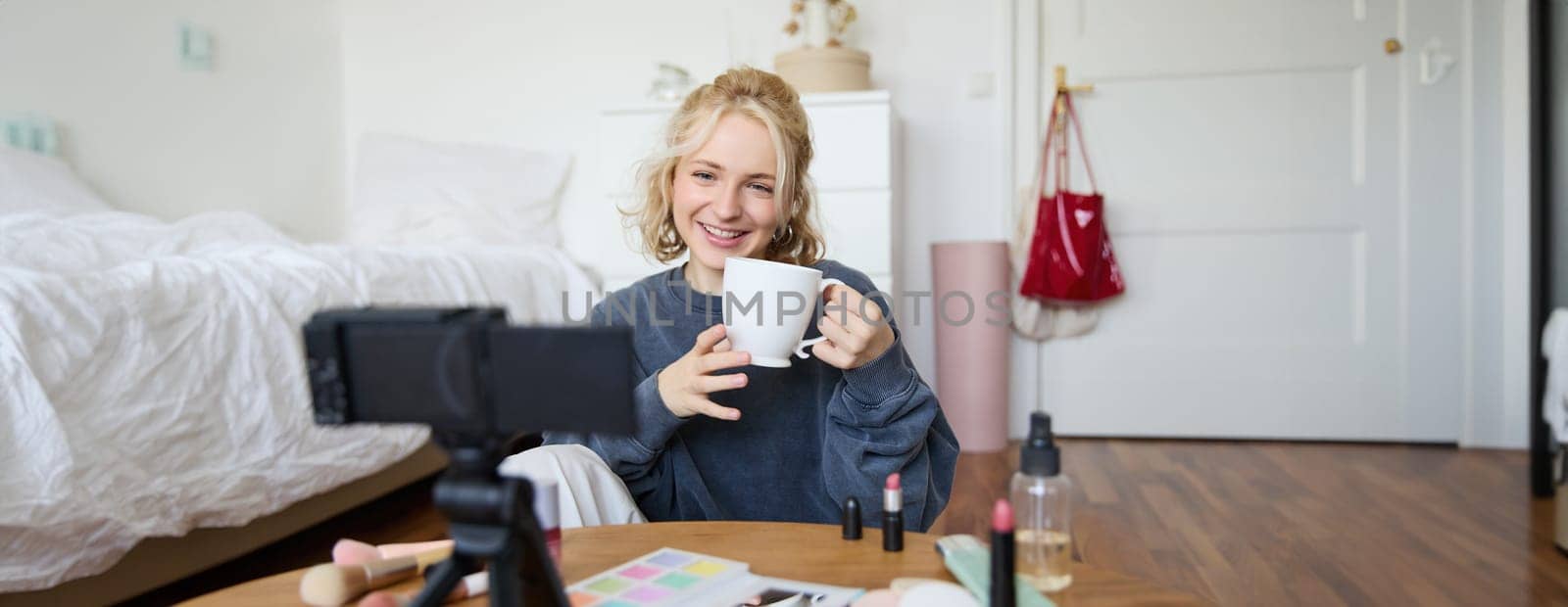 Cheerful woman, beauty blogger, records lifestyle vlog on digital camera, talks casually, tells a story for social media followers, holds cup, drinks tea and sits on floor in her room.