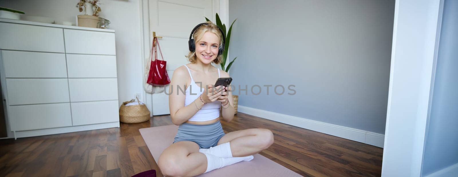 Portrait of young sporty woman in wireless headphones, sitting with smartphone on rubber mat, using workout training app, fitness application on her phone, listening in earphones.