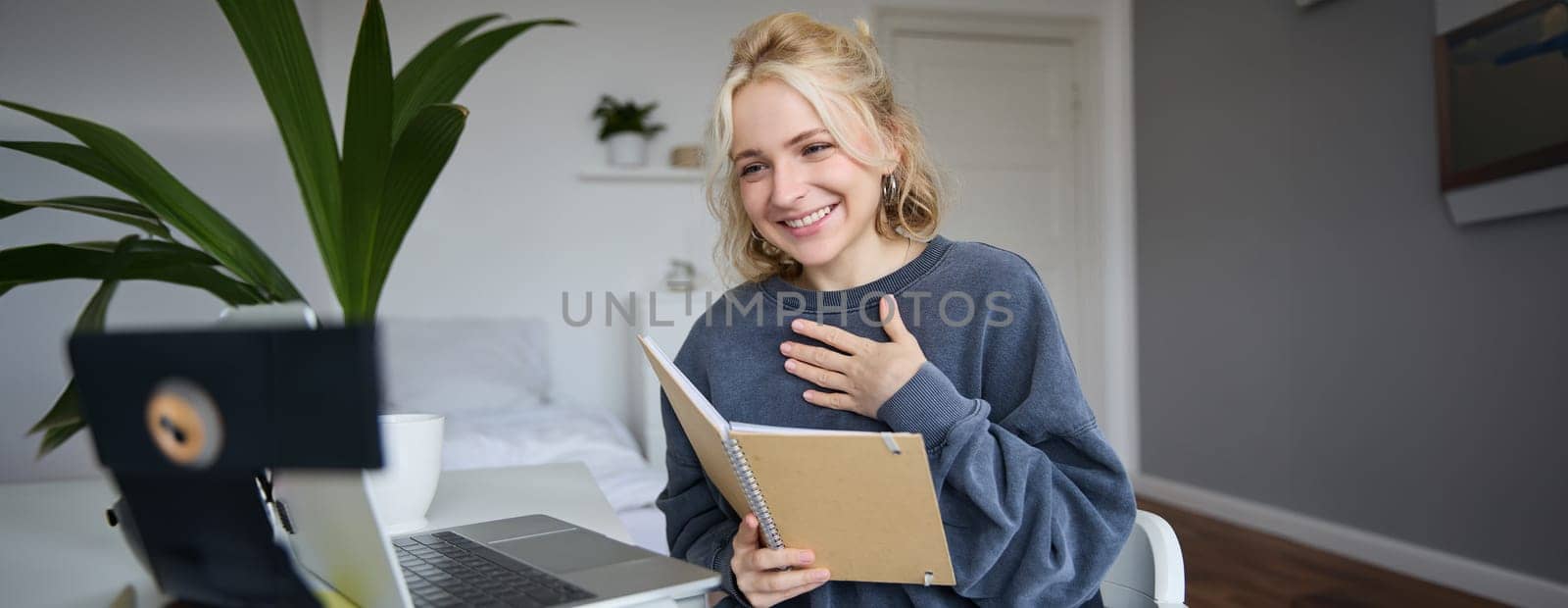 Portrait of young smiling blond woman, working from home, online chatting, using digital video camera, recording vlog, holding notebook, reading notes, explaining something by Benzoix