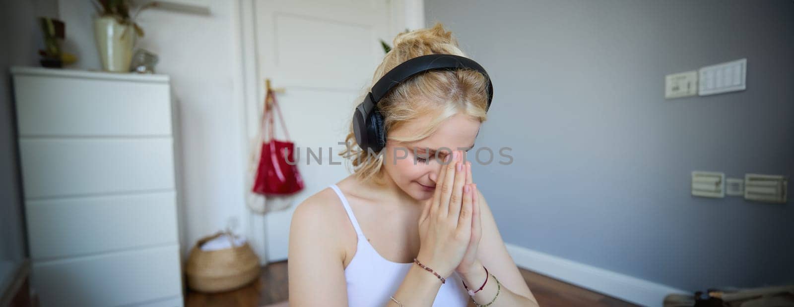 Close up portrait of young sporty woman in headphones, finish yoga training session, bowing with namaste hand sign, meditating by Benzoix