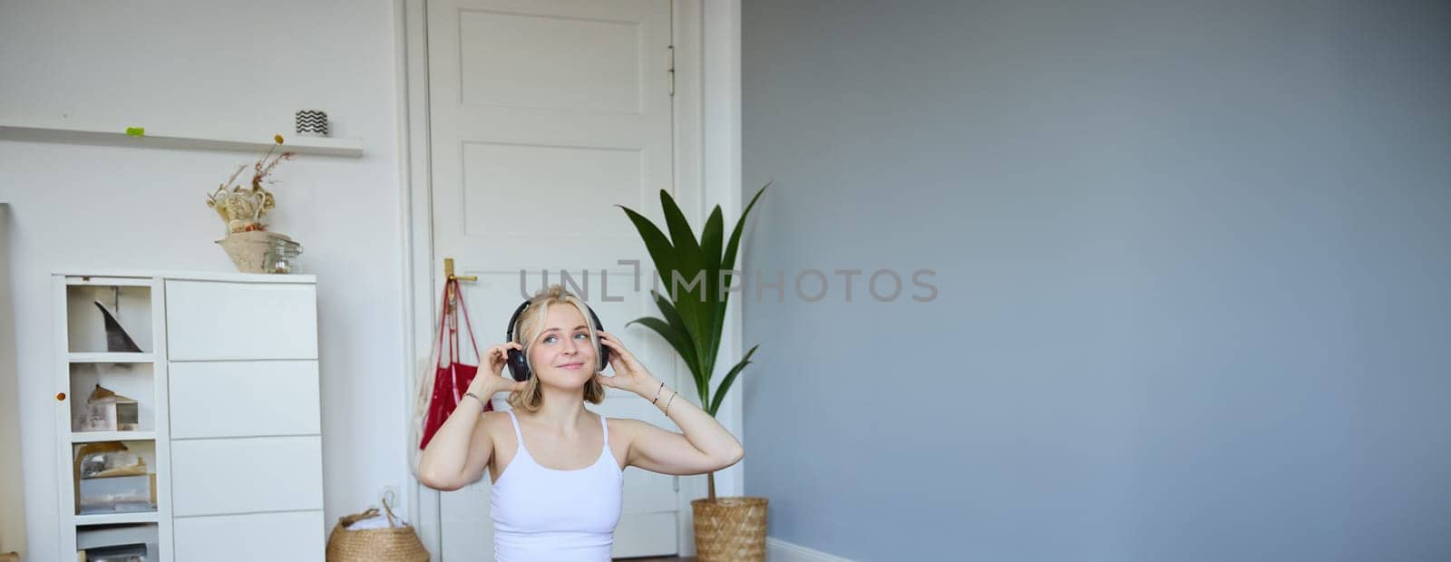 Vertical shot of young sporty woman doing workout at home, putting on headphones, watching video of fitness instructor on laptop, sitting on yoga mat.