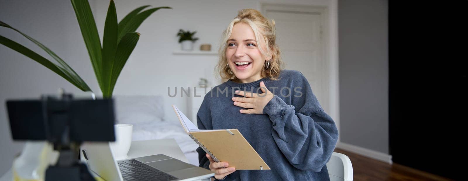 Portrait of smiling, beautiful young blond woman, student working on assignment from home, online learning in her bedroom, talking to video camera, chatting, holding notebook.