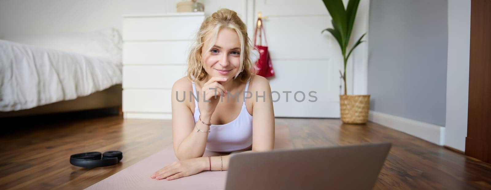Portrait of cute young fitness woman, doing workout at home, lying on rubber yoga mat, watching exercises online on laptop by Benzoix