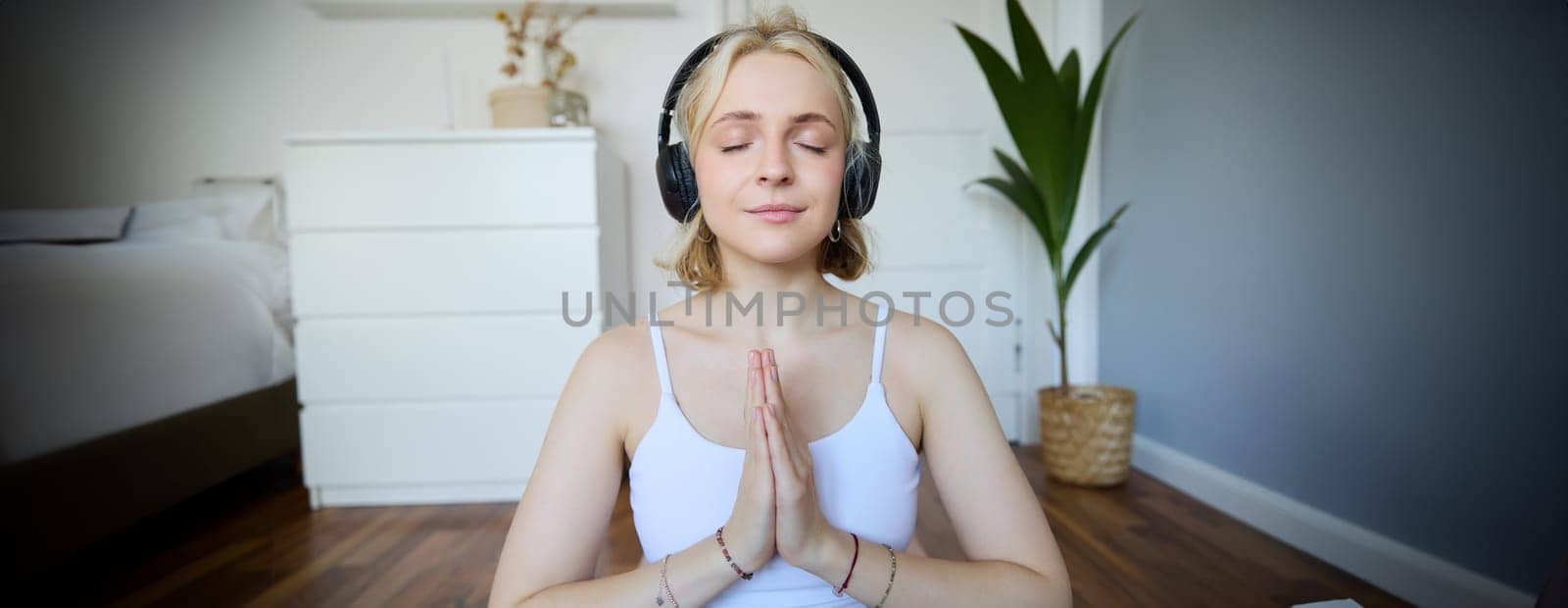 Portrait of woman meditating at home, sitting in headphones, listening to yoga podcast, holding hands in namaste sign by Benzoix