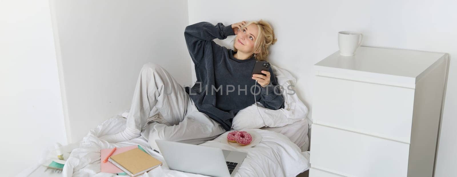 Lifestyle and weekend concept. Young smiling woman, relaxing at home, lying in bed with cup of tea and doughnut, using laptop and smartphone in bedroom.