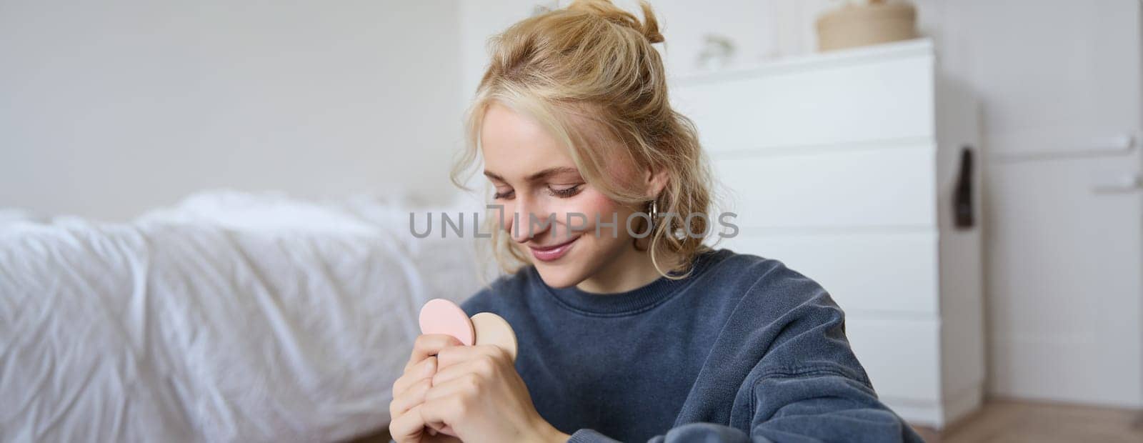 Image of young blond woman, girl records video about makeup, shows skin tone beauty products, sits in a room on floor.