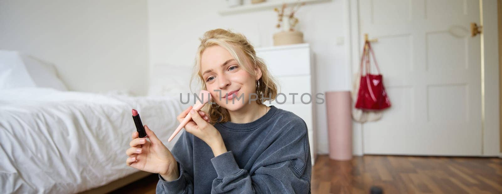 Woman beauty blogger, records video of herself sitting in a room and rating makeup products, puts on make up, holds lipstick and cosmetic brush in hand, using professional camera for content creation by Benzoix