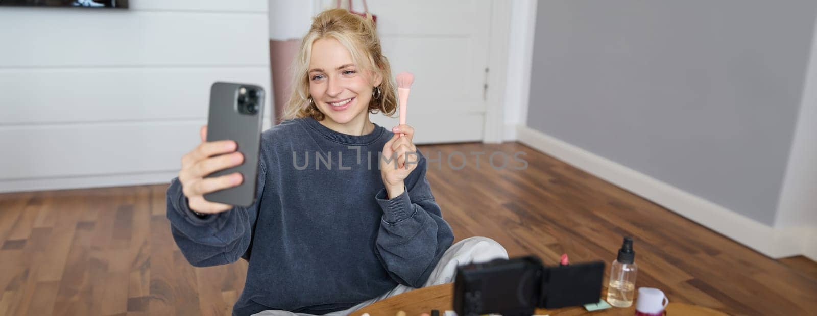 Image of stylish young woman, social media influencer, taking pictures on mobile phone, doing makeup tutorial for followers online, recording video vlog in her bedroom, showing brush by Benzoix