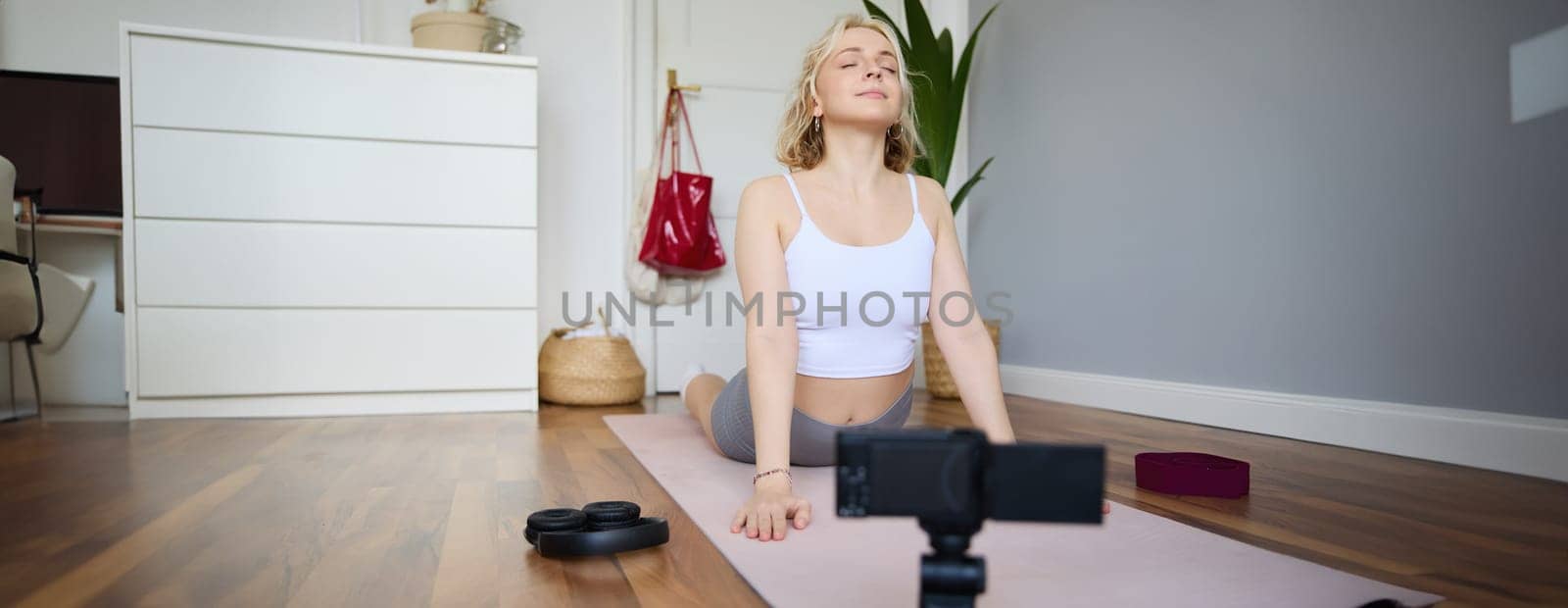 Portrait of young blogger, yoga content creator, showing exercises, recording video of herself working out at home on rubber mat.