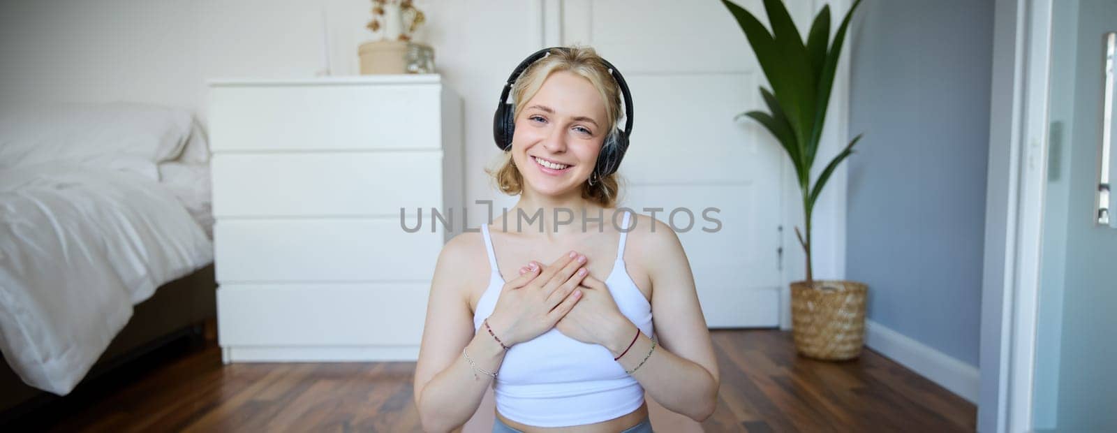Portrait of woman feeling relaxed and in peace after meditation or yoga training at home, holding hands on chest, wearing headphones, smiling at camera.