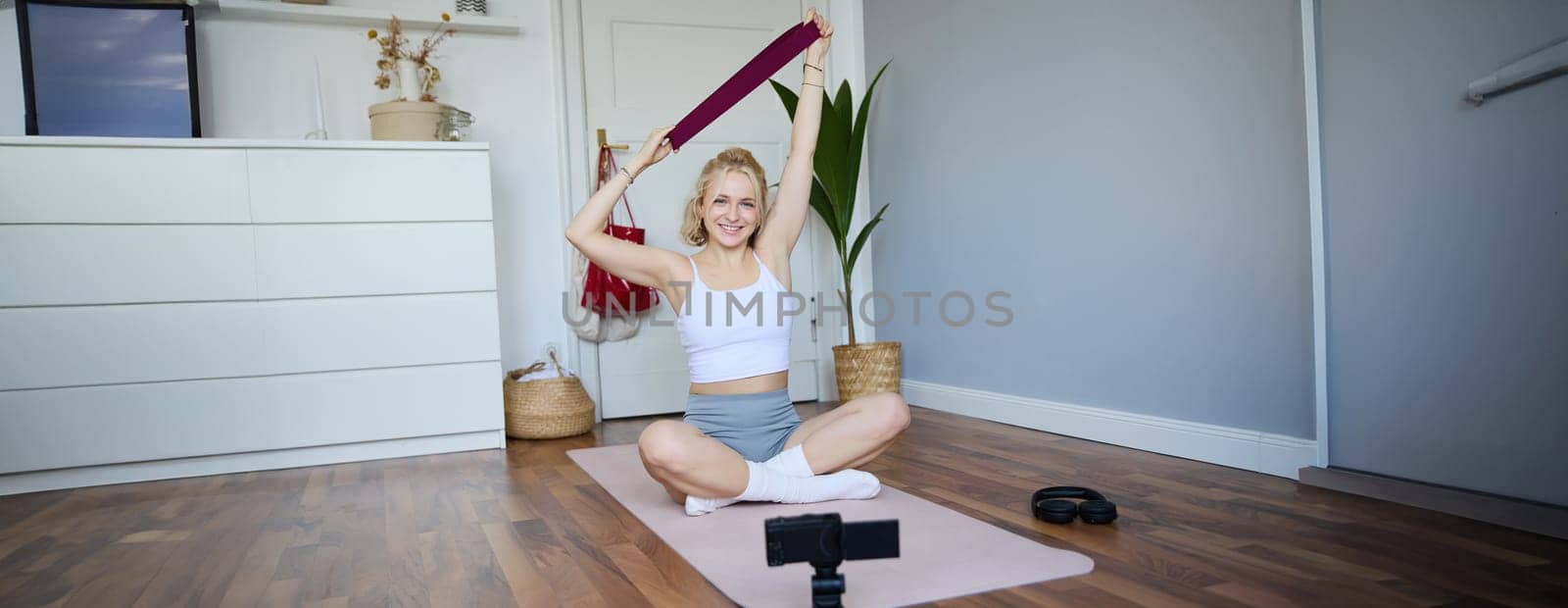 Image of smiling, athletic fitness instructor, female social media influencer, recording video about workout, showing how to use resistance bands on digital camera, sitting on rubber yoga mat at home by Benzoix