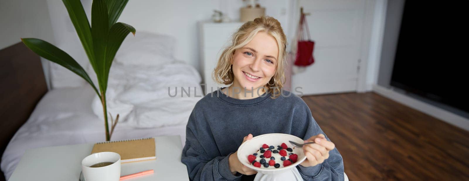 Cute girl eats breakfast and drinks tea in her room, makes herself healthy lunch in bowl, sits in bedroom.