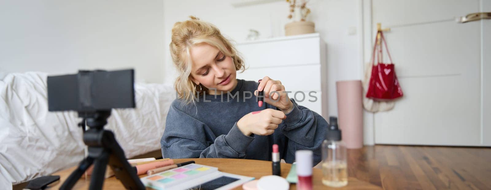 Woman beauty blogger, records video of herself sitting in a room and rating makeup products, puts on make up, holds lipstick and cosmetic brush in hand, using professional camera for content creation.