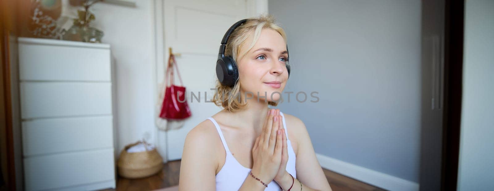 Close up portrait of young relaxed woman in headphones, holding hands together in namaste gesture, listening to meditation podcast, practice yoga by Benzoix