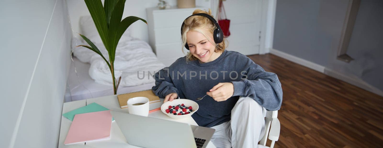 Portrait of smiling young blond woman in headphones, sitting in room, watching movie on laptop, eating breakfast and drinking tea, having lunch in front of computer screen.
