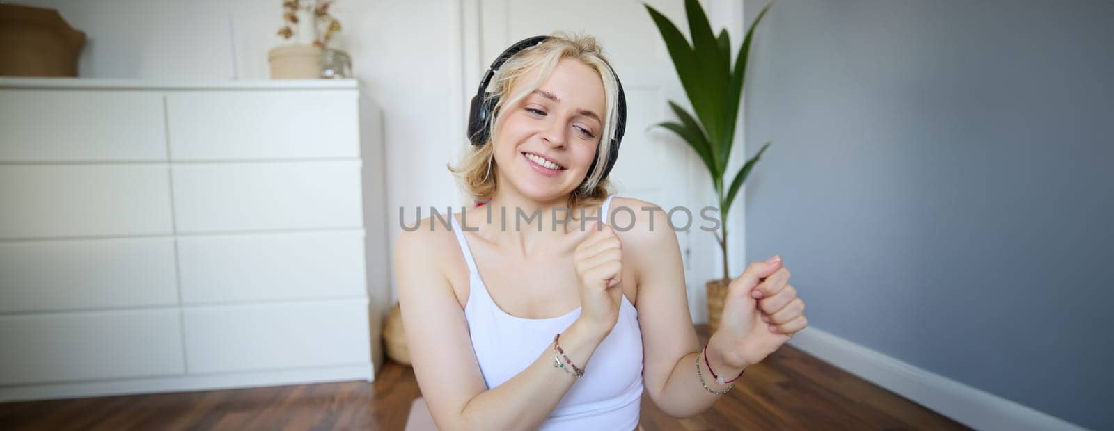 Portrait of happy, beautiful athletic woman, working out at home, listening music to boost energy while active training session, using wireless headphones and rubber yoga mat.