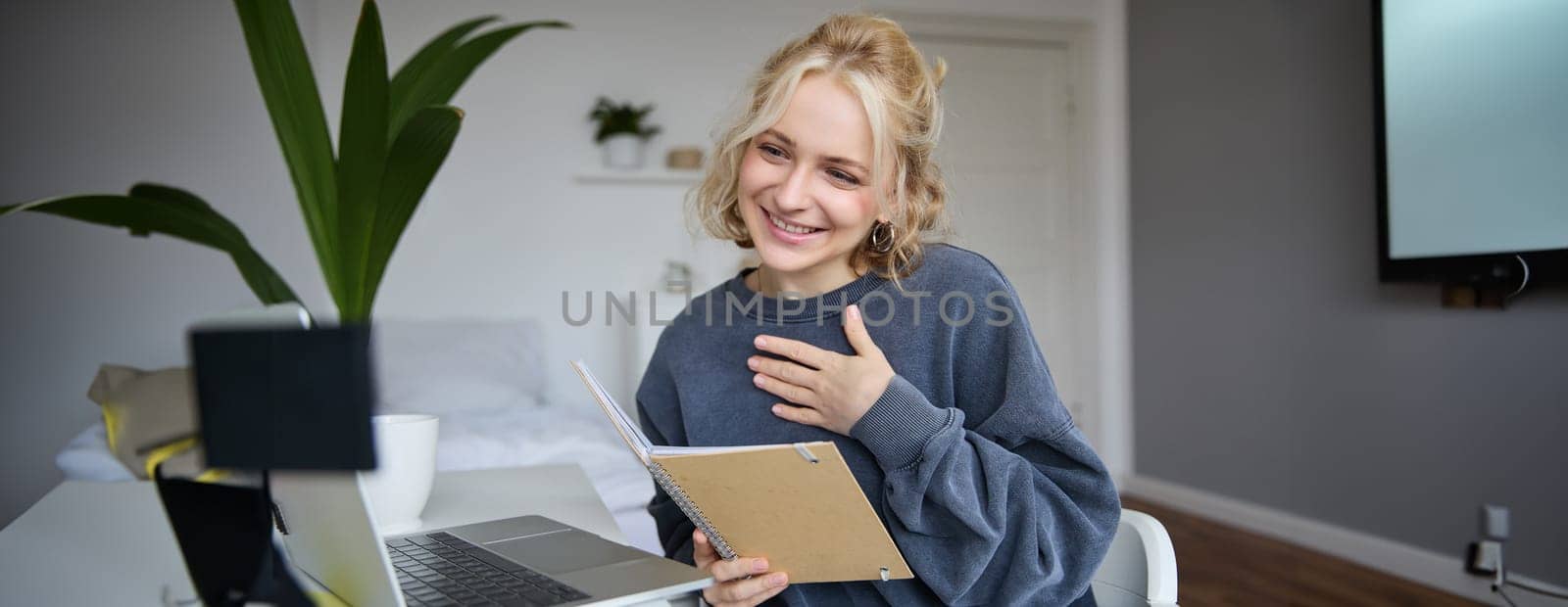 Portrait of smiling, beautiful young blond woman, student working on assignment from home, online learning in her bedroom, talking to video camera, chatting, holding notebook.