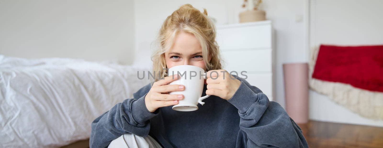 Portrait of young blond woman in casual clothes, sits on bedroom floor with cup of tea, drinking and looking at camera.