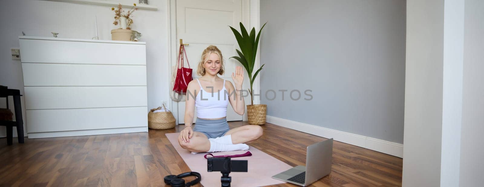Portrait of young female athlete, fitness trainer recording vlog, training session on digital camera, sitting in a room on rubber yoga mat, showing exercises.