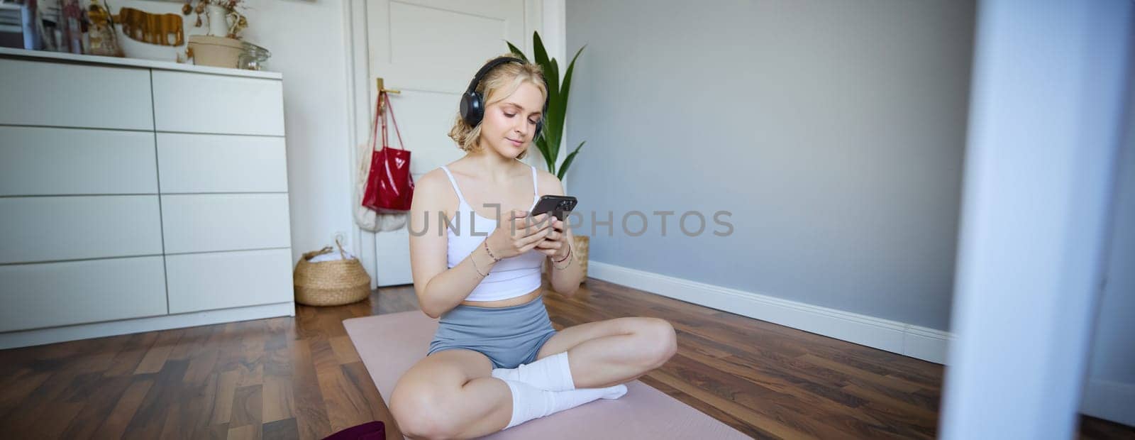 Portrait of young blond woman in headphones, turning on yoga, workout app on smartphone, choosing music on mobile phone application, sitting on rubber mat in room.