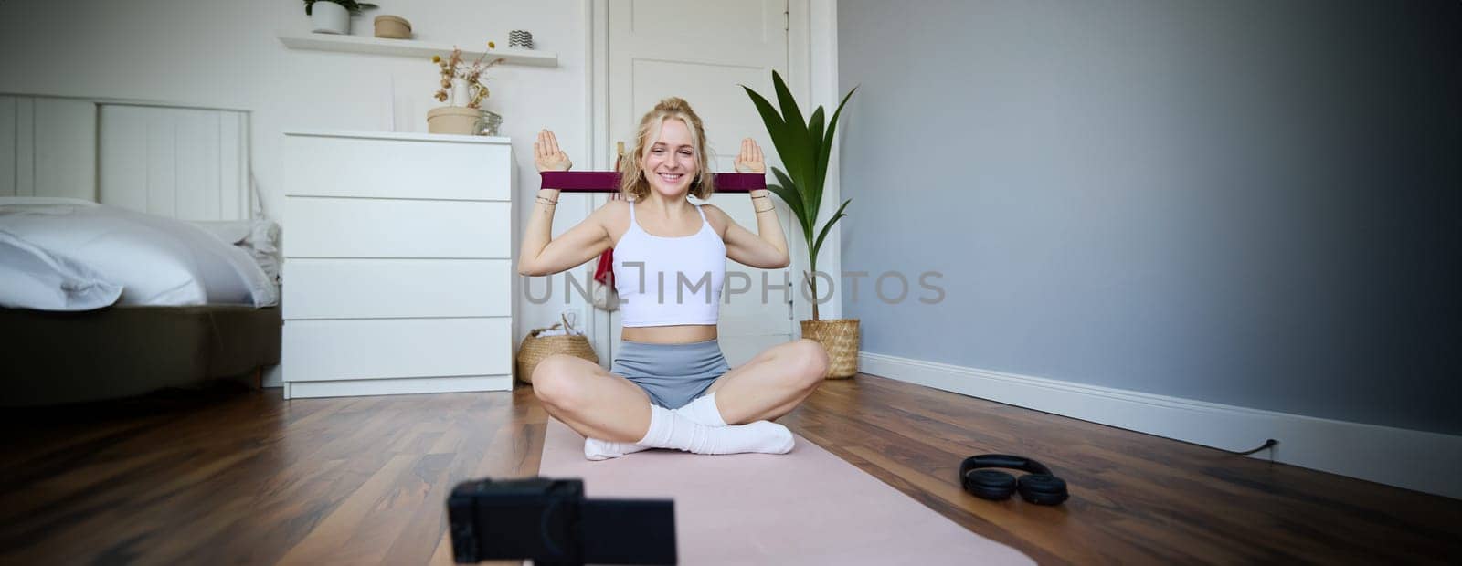 Portrait of young athletic woman recording home workout video, shooting content for sport fitness vlog, using resistance band and digital camera by Benzoix