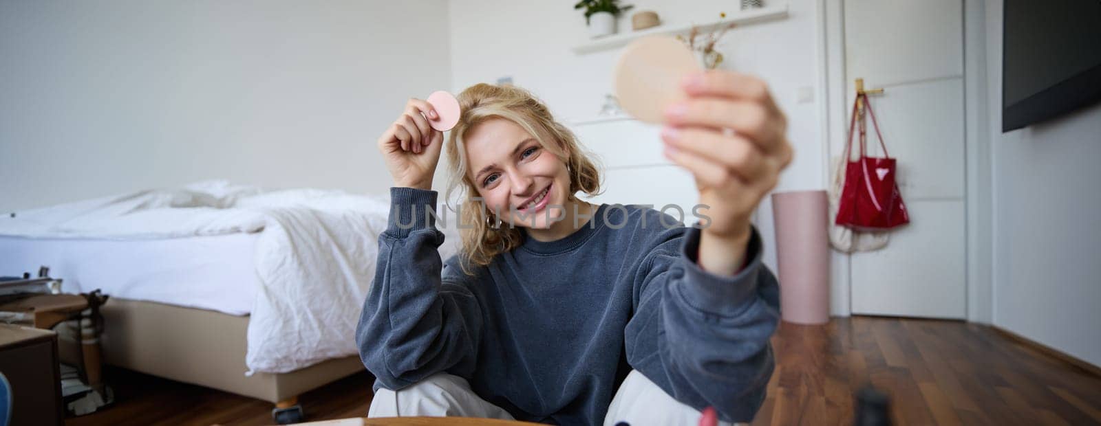 Portrait of young woman, content creator, showing beauty makeup products at camera, sitting on floor in bedroom and smiling.