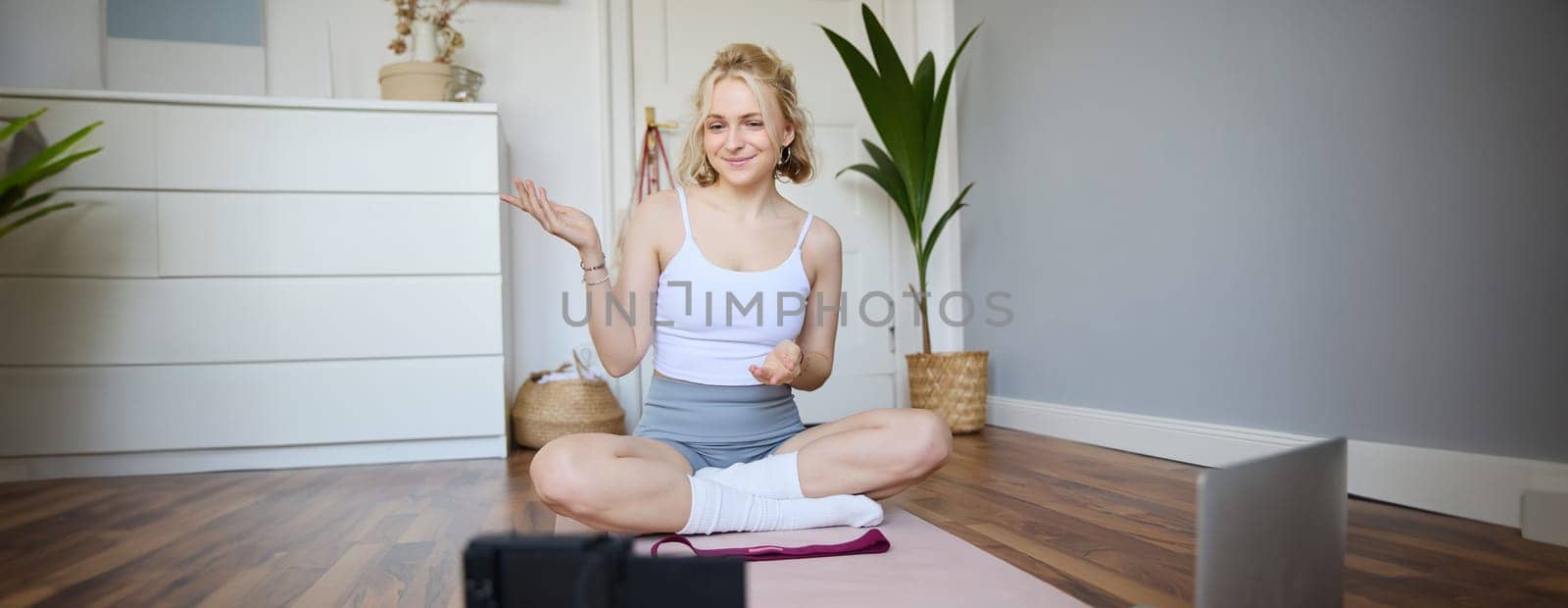 Portrait of beautiful fitness blogger, recording video on digital camera, showing workout exercises, explaining fitness movements or yoga training to followers, sits on rubber mat by Benzoix