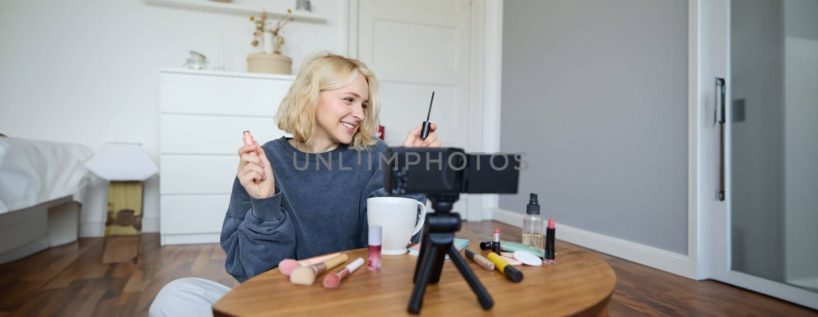 Portrait of young smiling woman in her room, recording video on camera, lifestyle vlog for social media, holding mascara, reviewing her makeup beauty products, showing how to use cosmetics by Benzoix