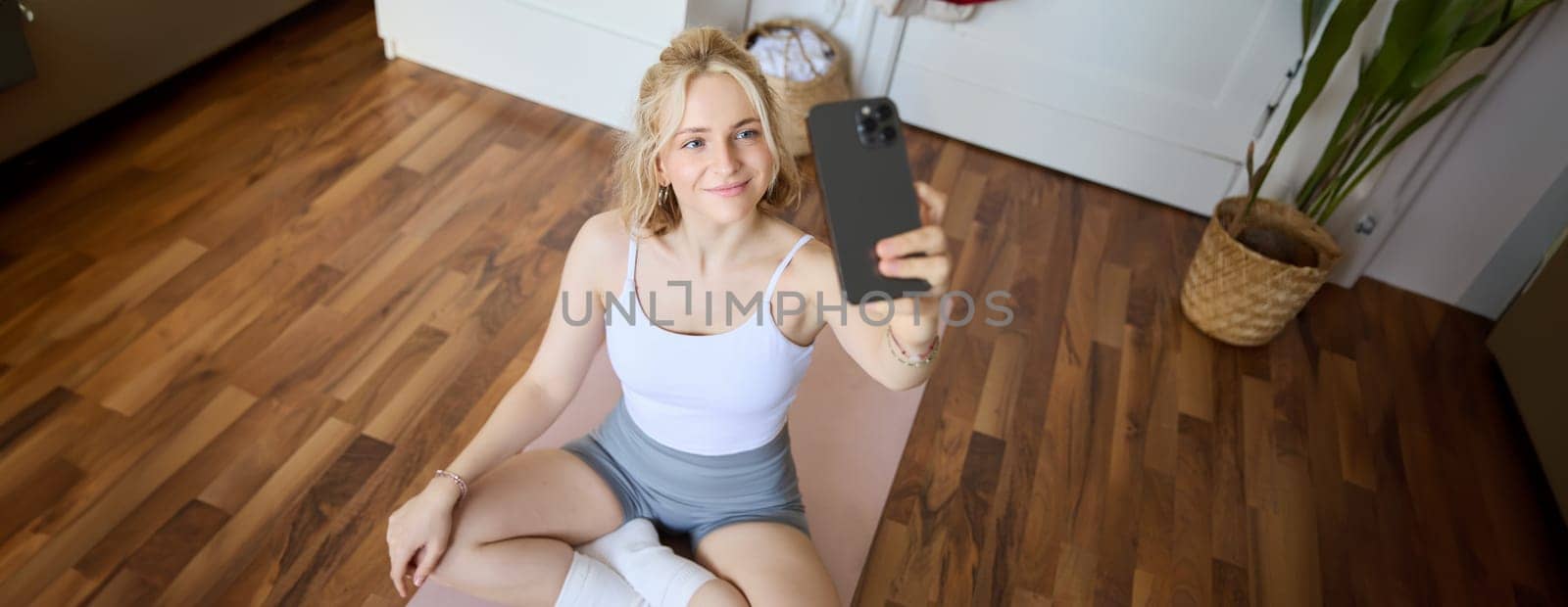 Portrait of stylish, young beautiful woman takes selfie while working out at home, using smartphone to make photos on yoga mat, posing in fitness clothes by Benzoix