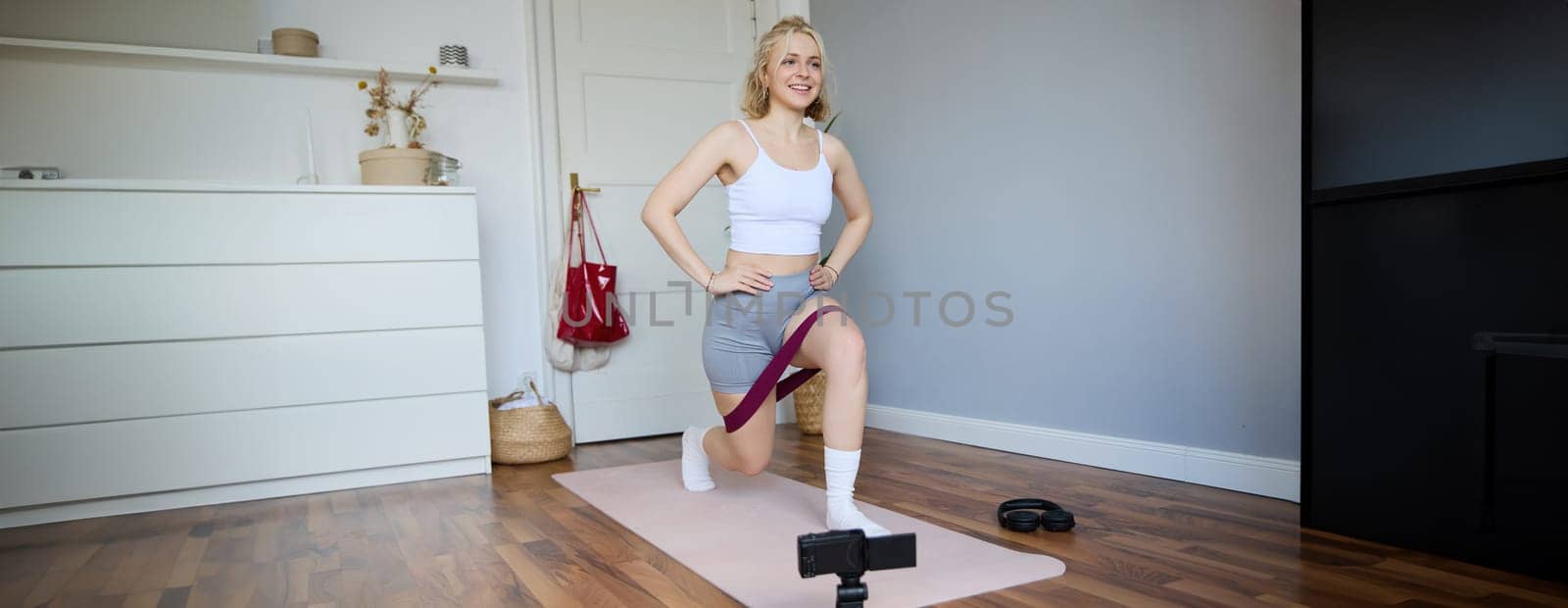 Portrait of fitness instructor, young sporty vlogger woman doing exercises on camera, shooting video about workout, using resistance band and doing sit-ups at home in empty room by Benzoix