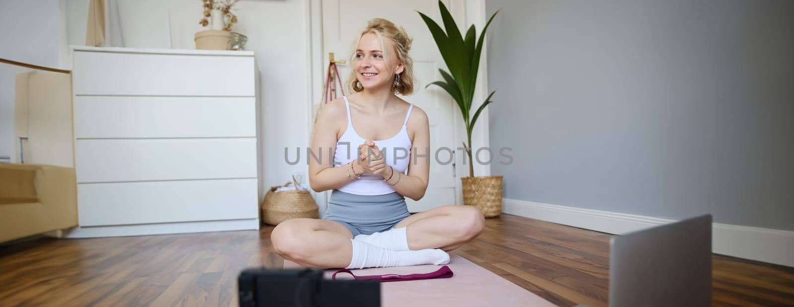 Portrait of young fitness instructor, vlogger showing exercises on camera, recording herself, sitting on mat with laptop, doing workout, explaining yoga movements to followers by Benzoix