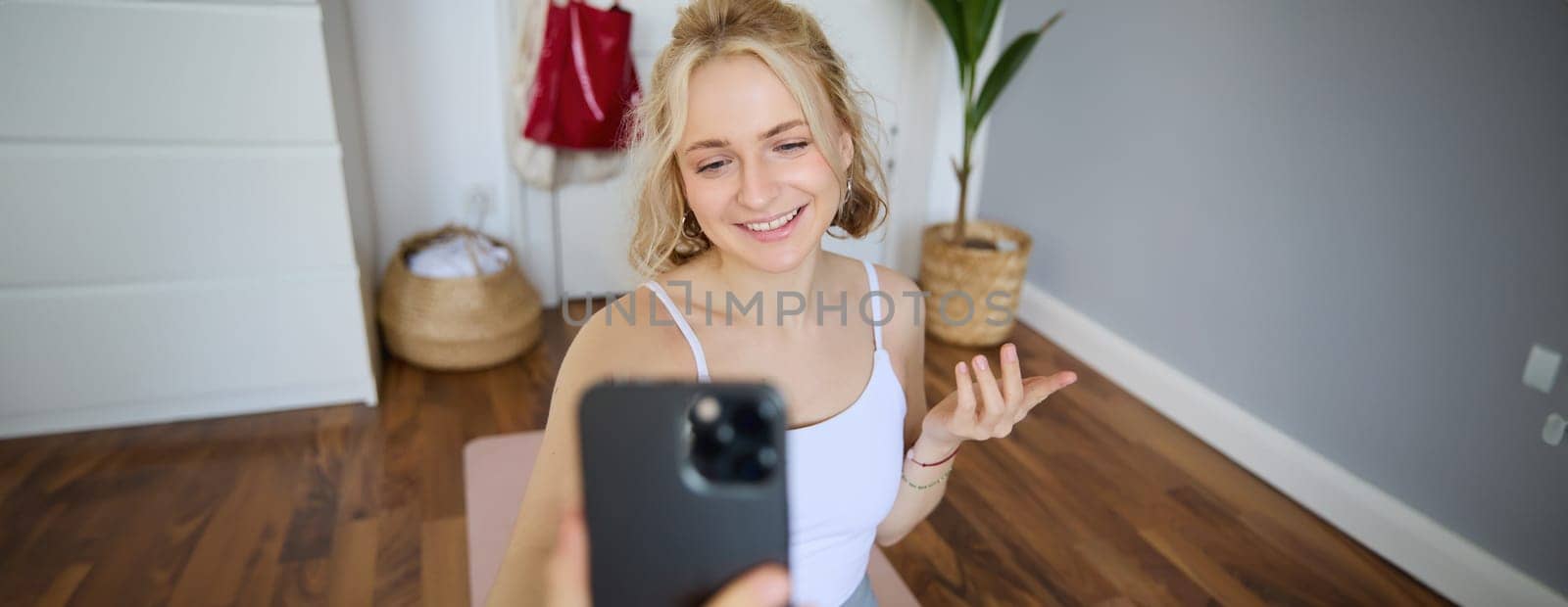Portrait of beautiful young fitness trainer, showing home workout, taking selfie on mobile phone while sitting on yoga rubber mat in bedroom, wearing activewear by Benzoix